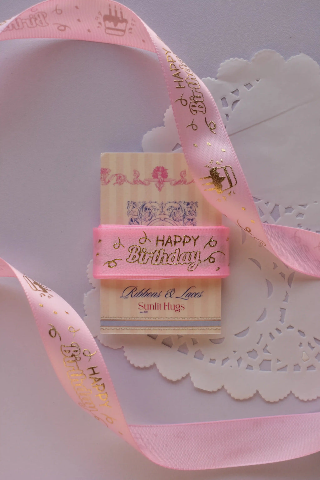 Soft pink birthday ribbon with gold foil script wrapped straight across a "Sunlit Hugs" card, resting on a white lace doily.