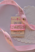 Soft pink birthday ribbon with gold foil script wrapped straight across a "Sunlit Hugs" card, resting on a white lace doily.