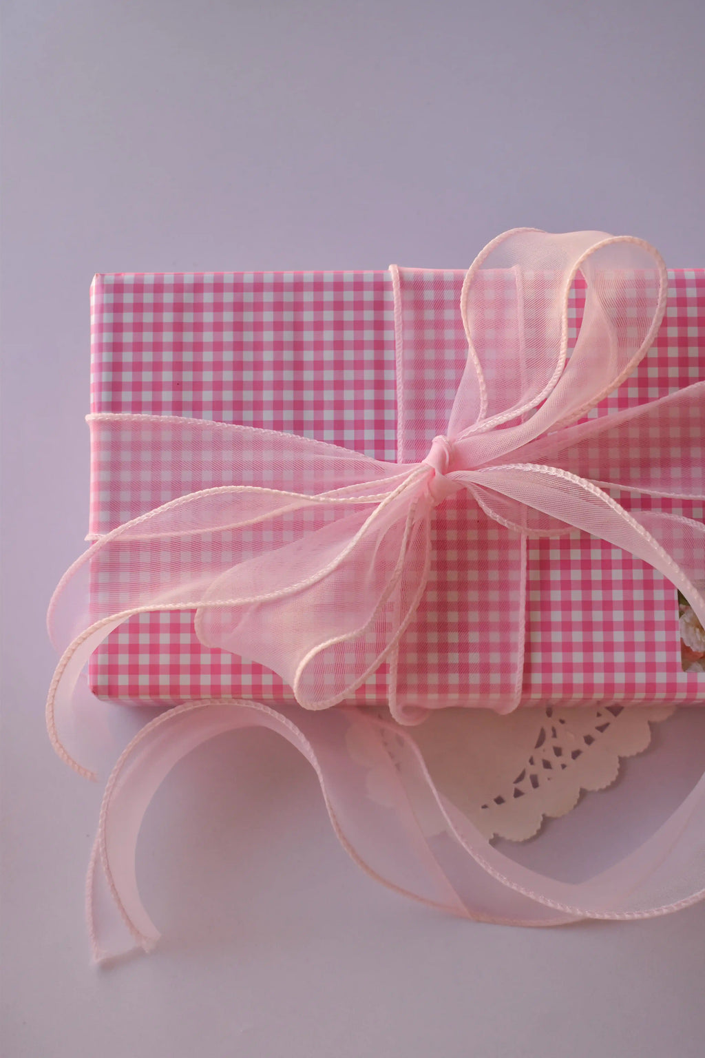 A gift wrapped in pink gingham paper, tied with a large sheer pink organza ribbon bow with long, flowing strands.
