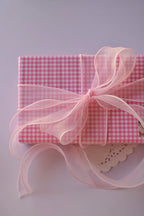 A gift wrapped in pink gingham paper, tied with a large sheer pink organza ribbon bow with long, flowing strands.