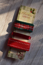A set of three vintage Christmas ribbons (green plaid, thin red satin, and wide red stitched grosgrain) on a festive postal-themed card stock label, on a light wooden surface with sunlight.