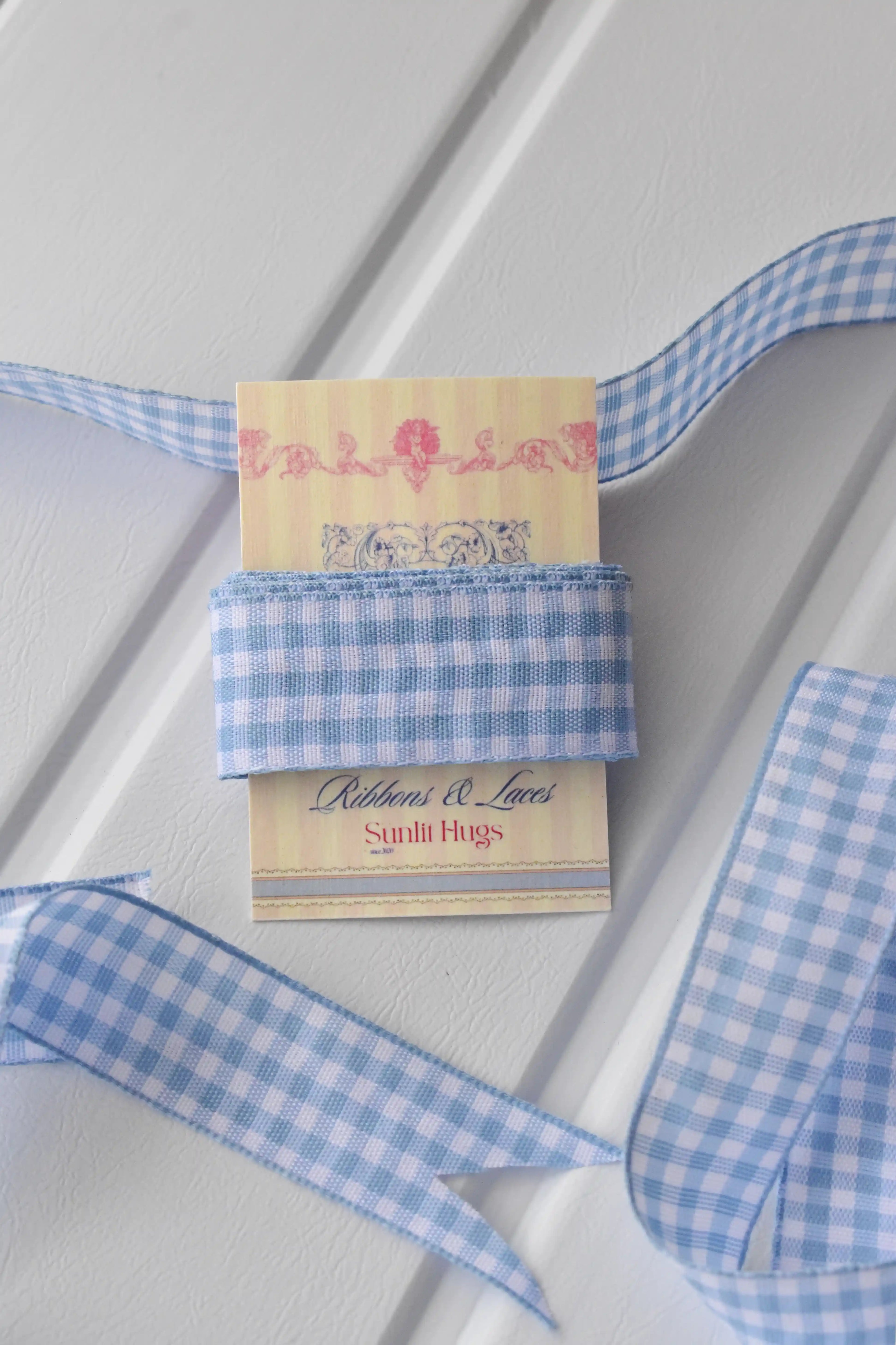 This image displays the Sky Gingham ribbon, showcasing its beautiful light blue and white checkered design and woven texture. A long strand is loosely unravelled to highlight the full design, with a section wrapped around a "Sunlit Hugs" brand tag.