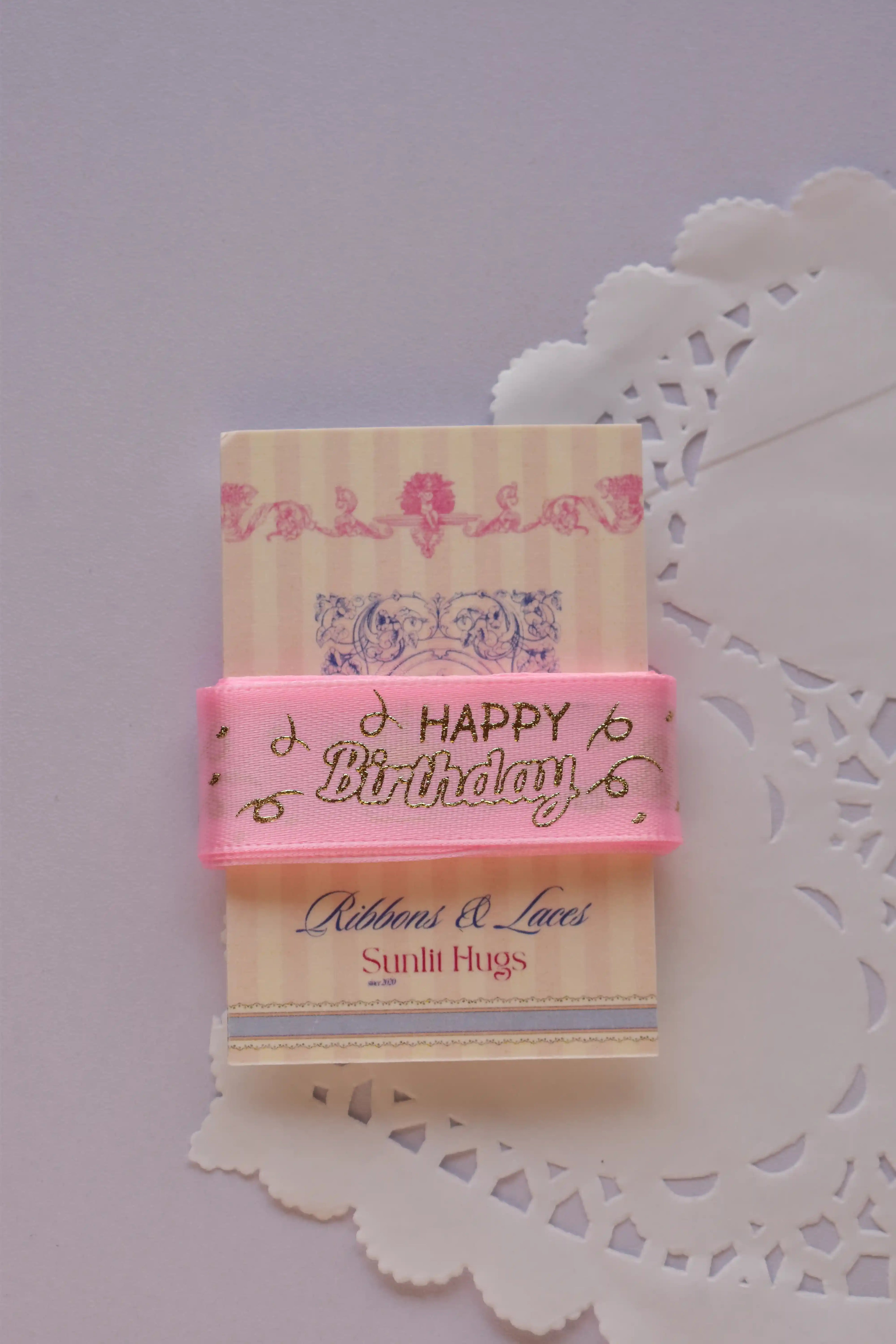 Soft pink satin ribbon with repeating gold foil "Happy Birthday" script draped around a "Sunlit Hugs" card on a lace doily.