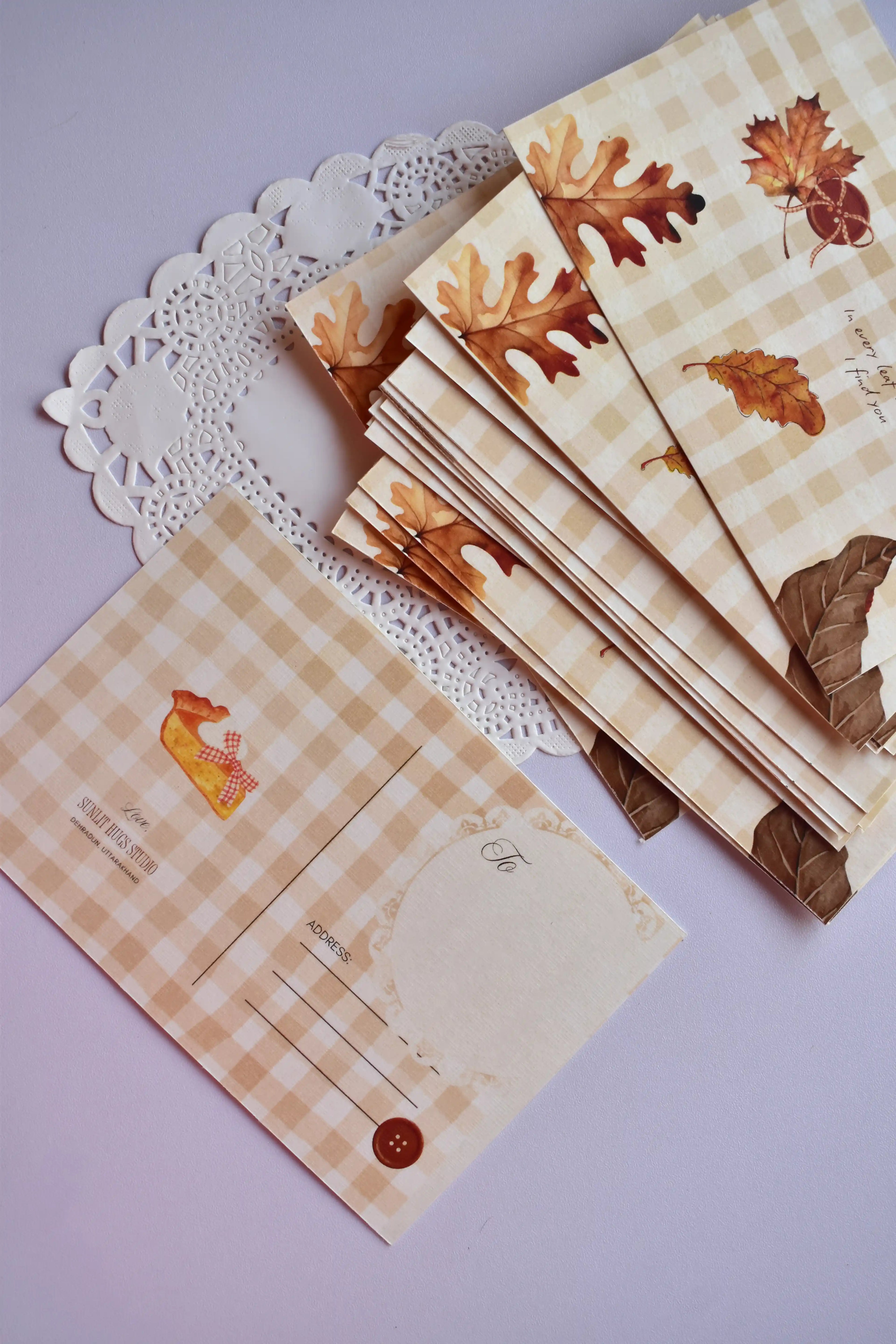 A stack of autumn postcards showing the front design of scattered brown and amber leaves and the emotional text. One postcard is turned over, displaying the pie slice and the address lines against the tan and cream gingham.

