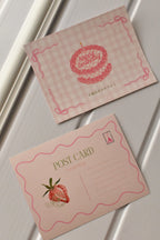 This image showcases the complete design of the Strawberry Birthday Postcard, featuring the pink gingham cake on the front and the pink strawberry illustration on the reverse side.