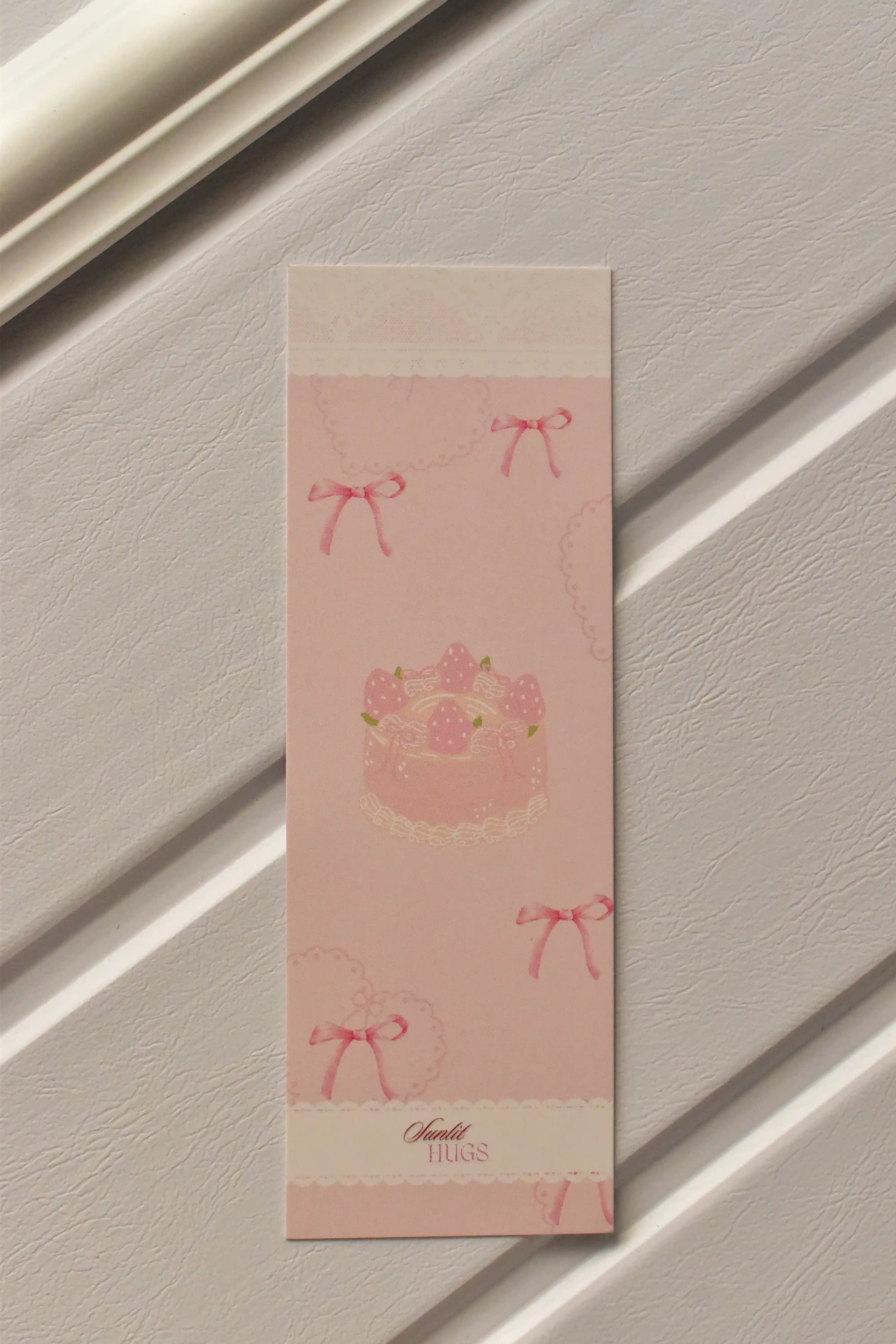 A detailed view of one of the three bookmarks, highlighting the charming pastel pink colour and sweet strawberry cake design with delicate bows, perfect for inspiring a cosy reading moment.