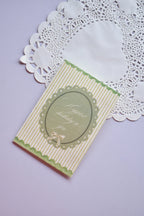 Close-up of a vintage-style greeting card with thin vertical pale green and white stripes. The card has a scalloped green border and a central oval label with a delicate ribbon bow, reading "Happiest birthday to you." It rests on a white lace paper doily.