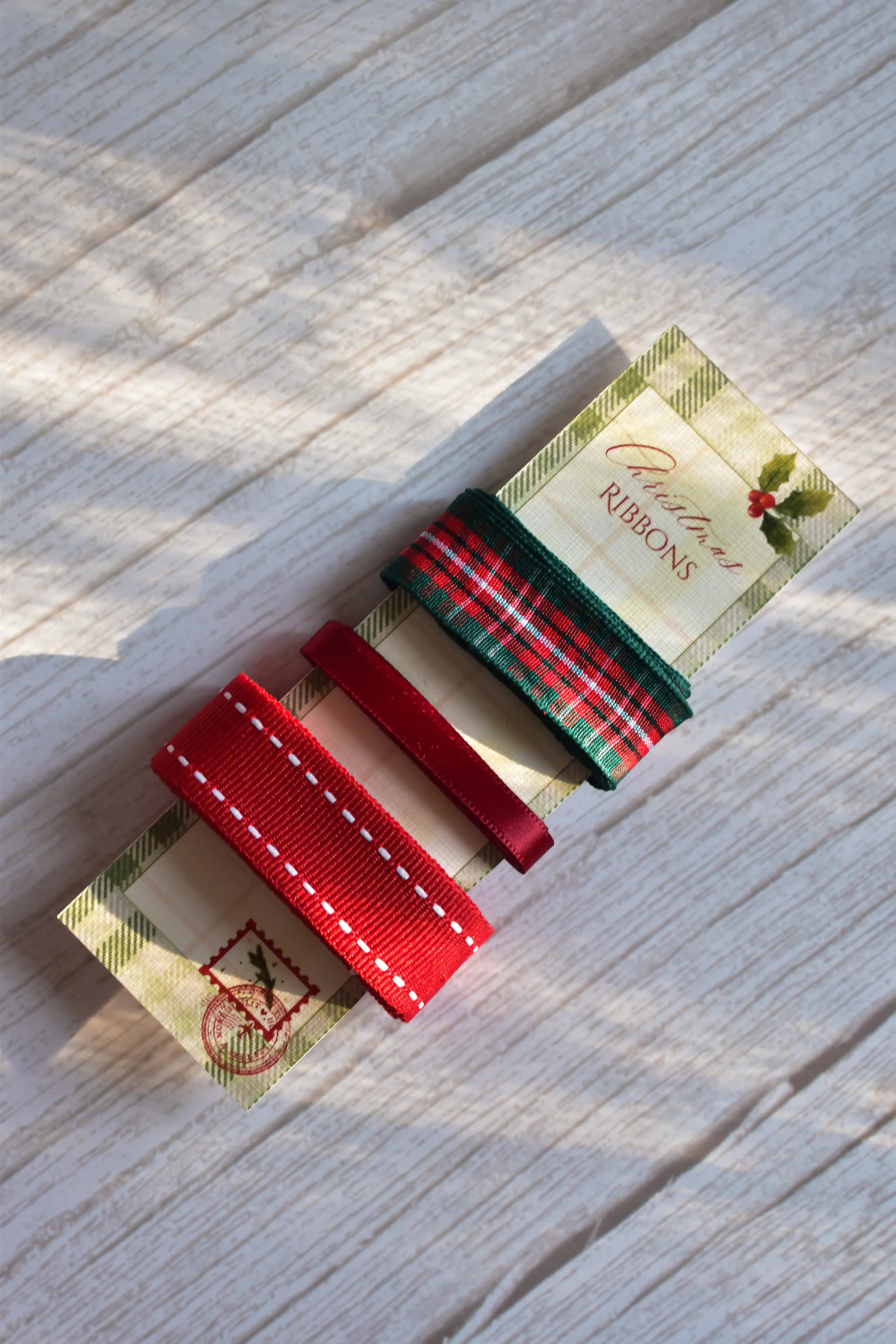 An angled view of the three Christmas ribbons (green plaid, thin red satin, and wide red stitched grosgrain) on their festive card stock label.