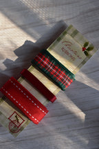 Close-up of three Christmas ribbons (plaid, thin satin, and wide stitched grosgrain) on their festive card stock label, with dramatic sunlight.