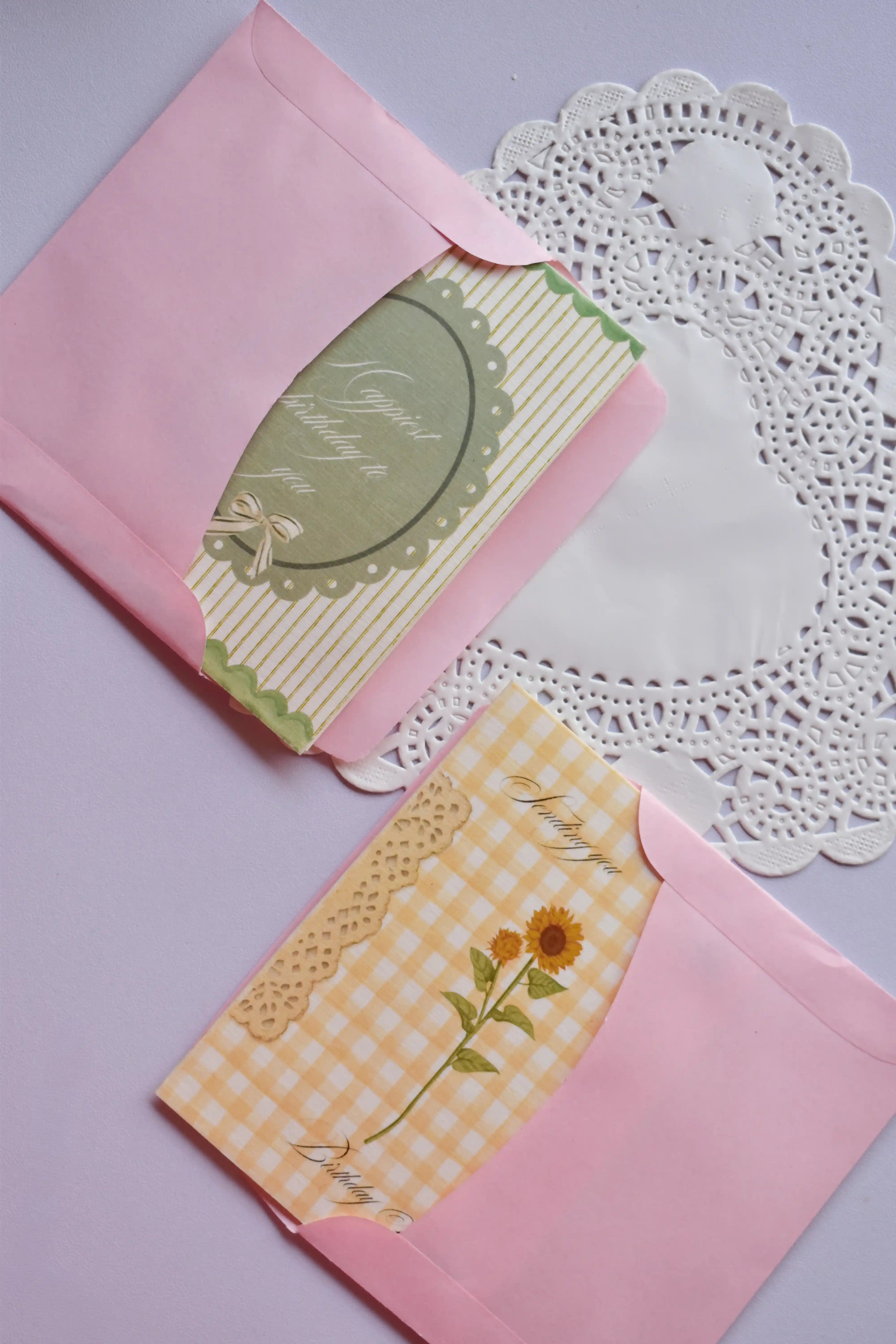 Two different vintage-style greeting cards partially tucked into two separate pink envelopes, both resting on a white lace paper doily. The top card is green and white striped, and the bottom card is yellow gingham with a sunflower stem.