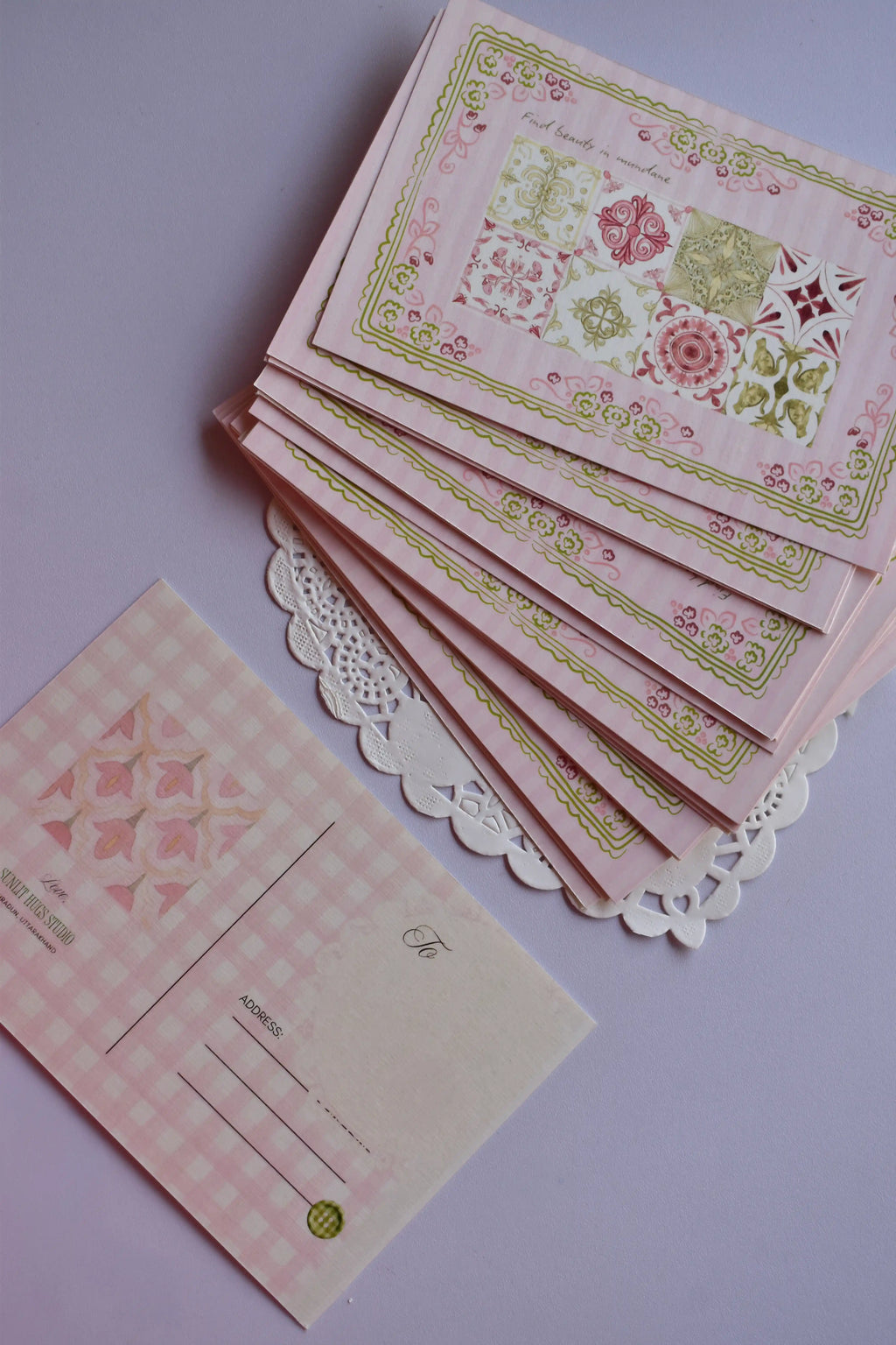 A neat stack of postcards showing the full front design: the nine-tile pattern and the 'Find beauty in mundane' quote. One card is flipped over, showing the pink gingham pattern of the reverse side.