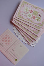 A neat stack of postcards showing the full front design: the nine-tile pattern and the 'Find beauty in mundane' quote. One card is flipped over, showing the pink gingham pattern of the reverse side.