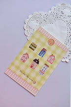 A vertical postcard displaying several colorful miniature homes and cottages against a pale yellow and green wide gingham background. The pink gingham scalloped border frames the phrase "You feel like home."