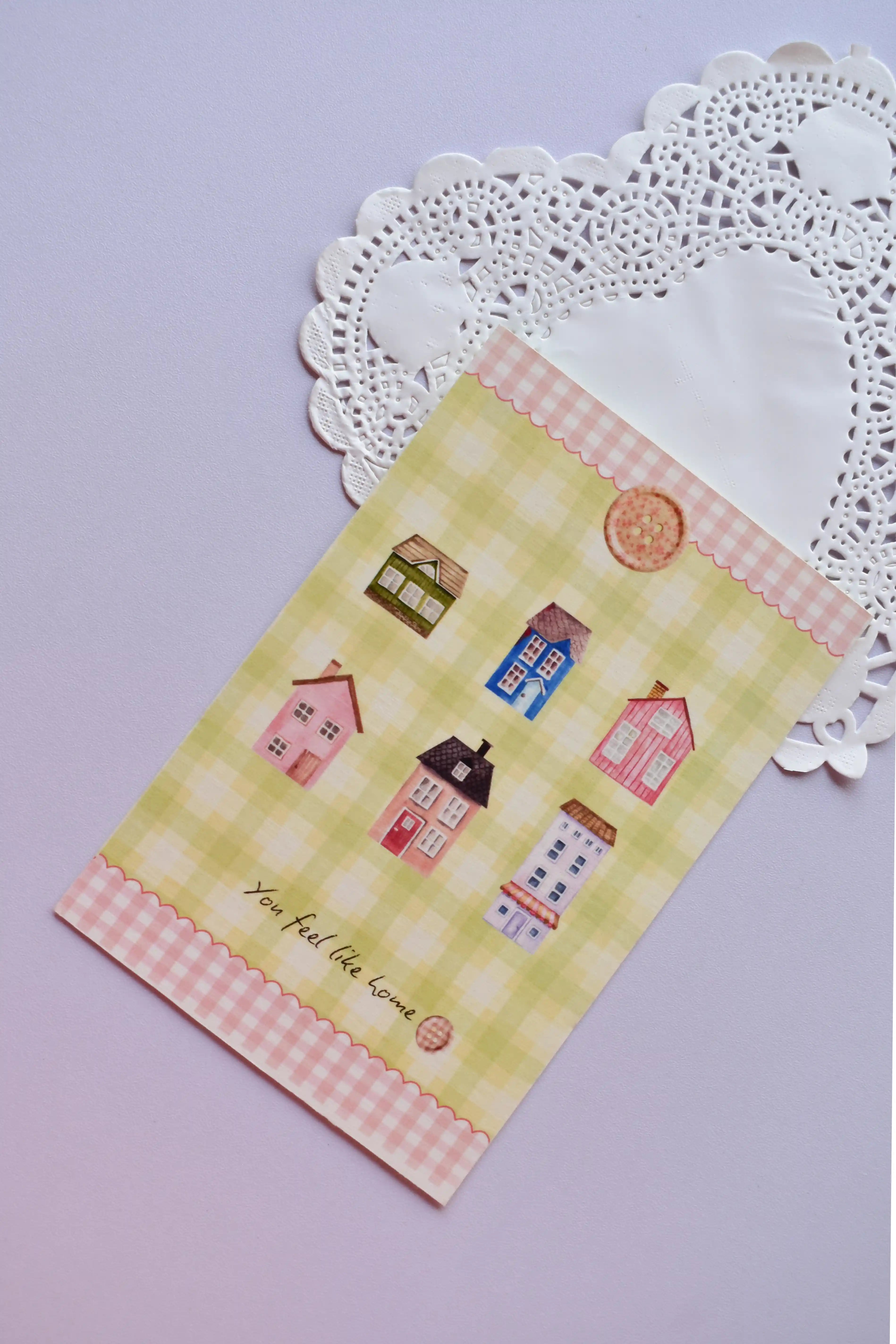 A vertical postcard displaying several colorful miniature homes and cottages against a pale yellow and green wide gingham background. The pink gingham scalloped border frames the phrase "You feel like home."