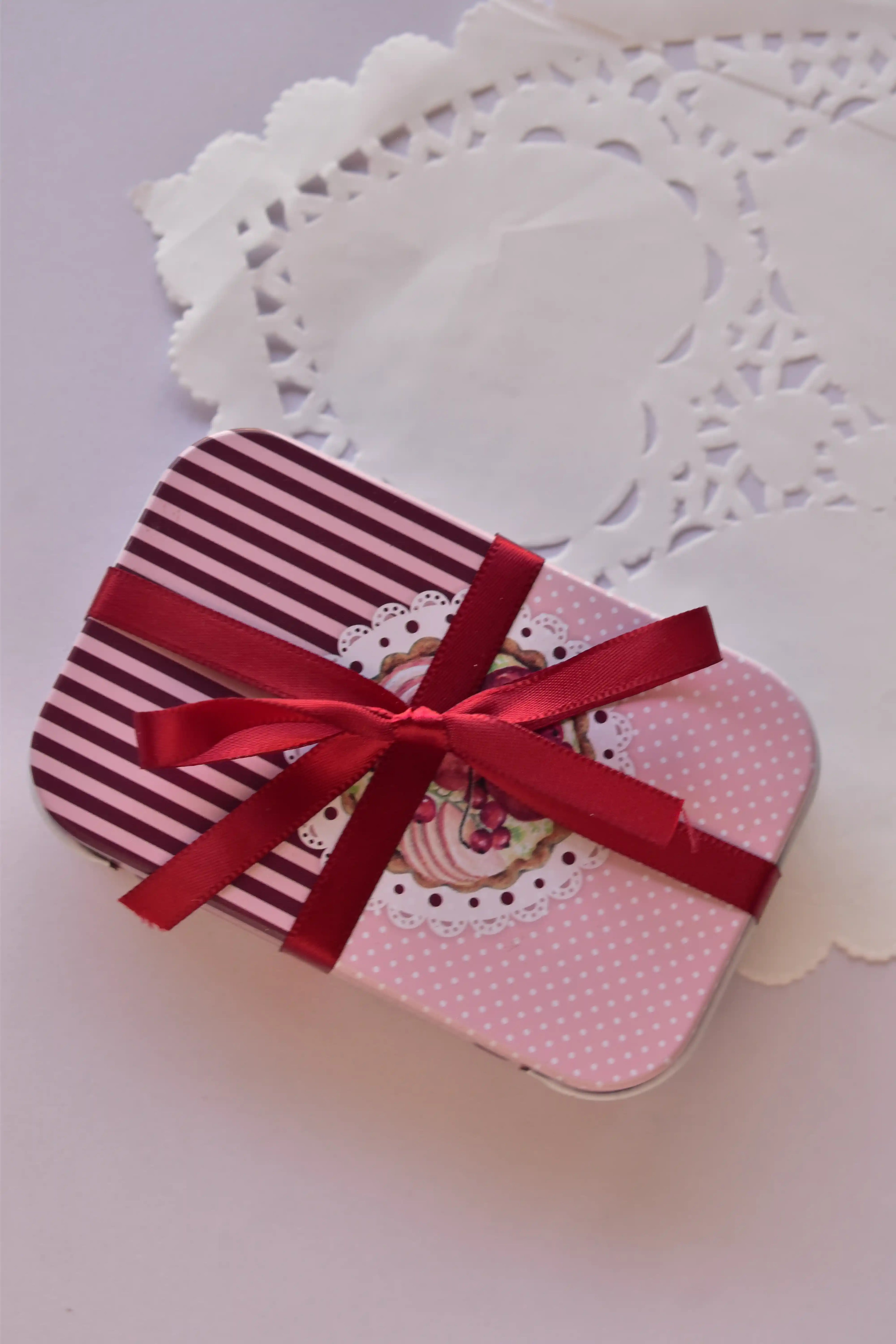A small pink and burgundy striped gift tin tied with a generous bow of narrow cherry red satin ribbon.